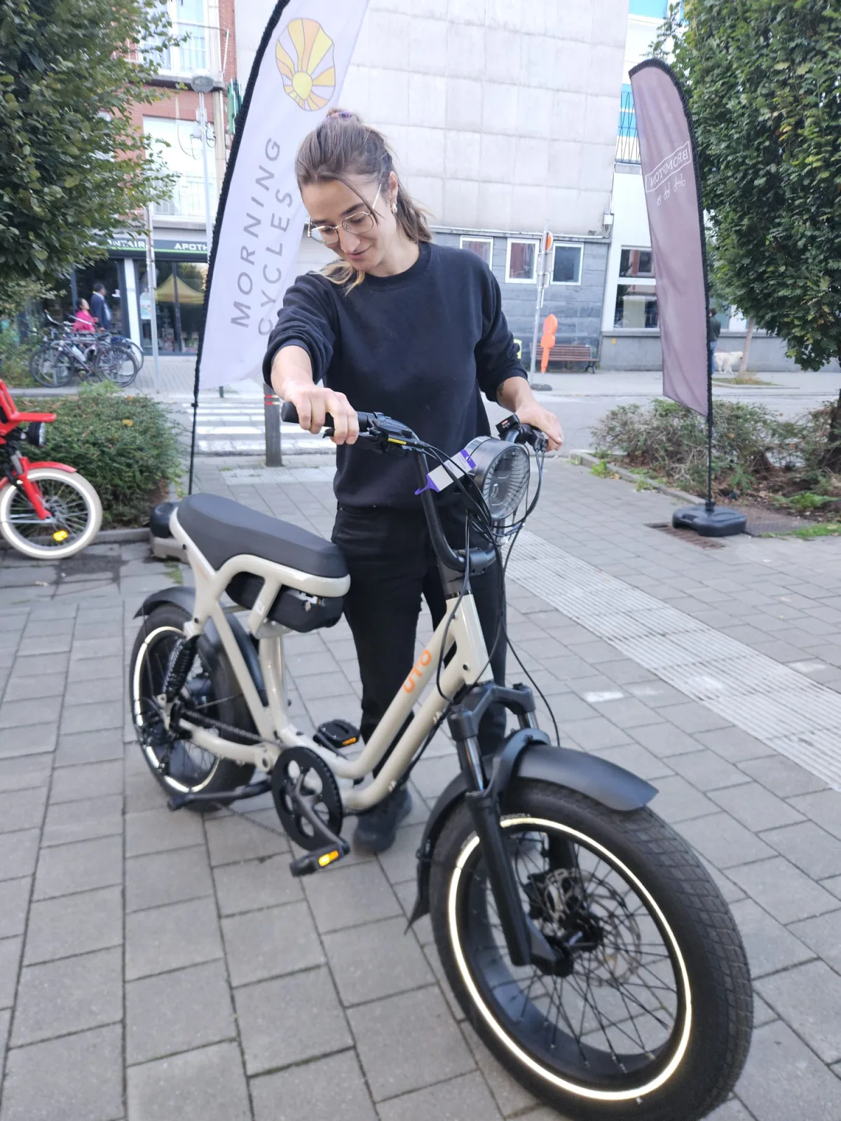 Test d'un vélo électrique devant le magasin Morning Cycles, à côté d'un drapeau Morning Cycles.