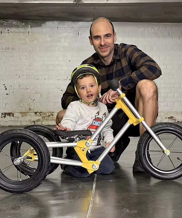 Jeune enfant casqué sur un tricycle Bonob jaune, un adulte accroupi à côté de lui