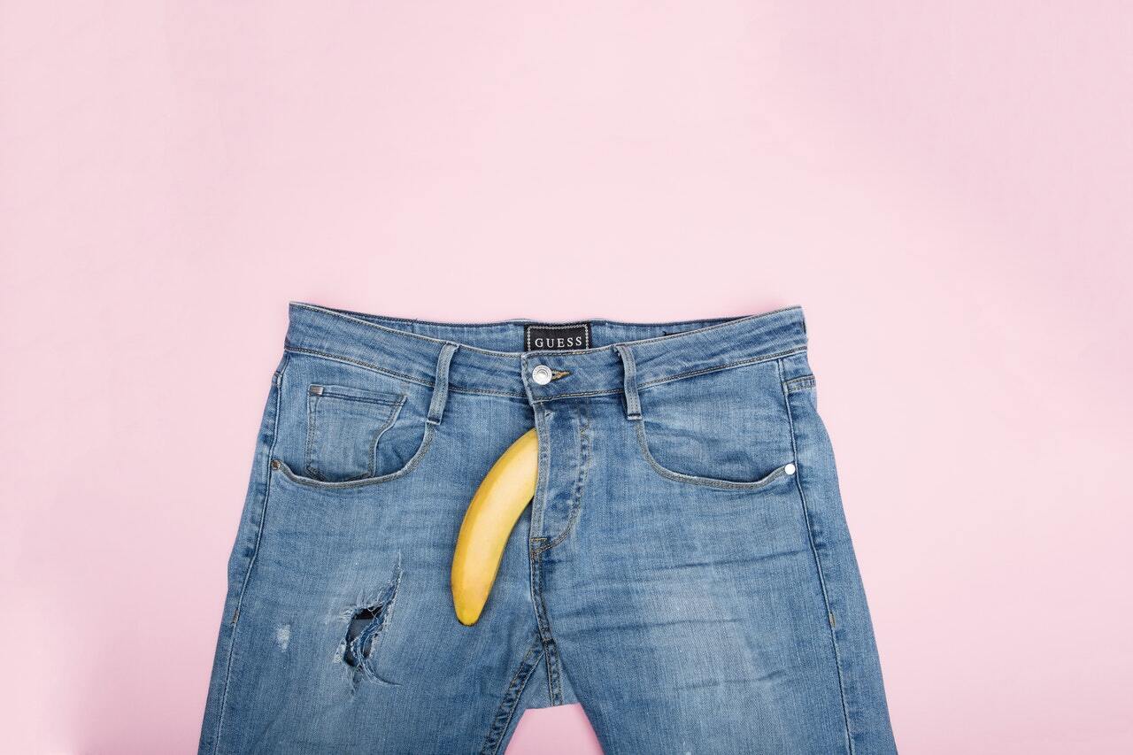 A banana is placed in the crotch area of a pair of jeans and is pointed downwards.