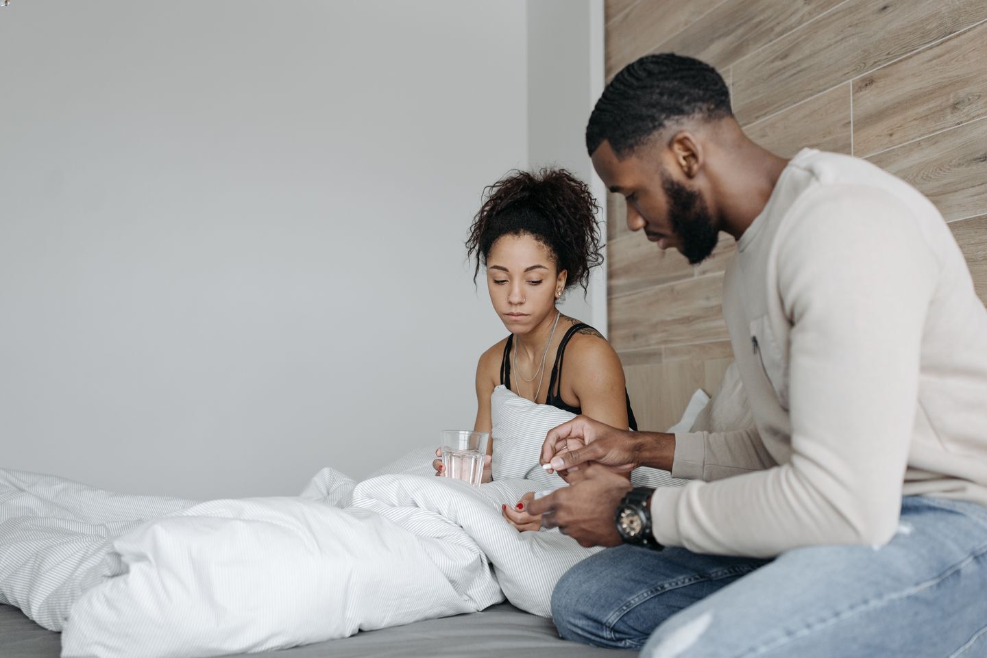 A couple sitting on a bed with the man holding a pill while the woman is holding a glass of water