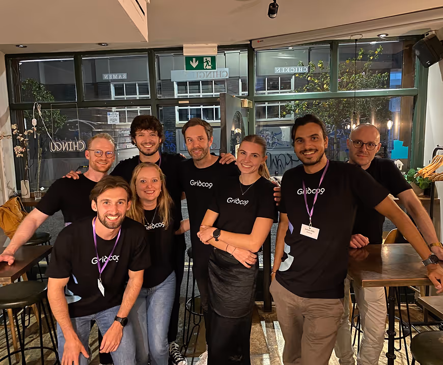 Group of eight smiling people wearing black Gridcap t-shirts posing together inside a restaurant with large windows at night.