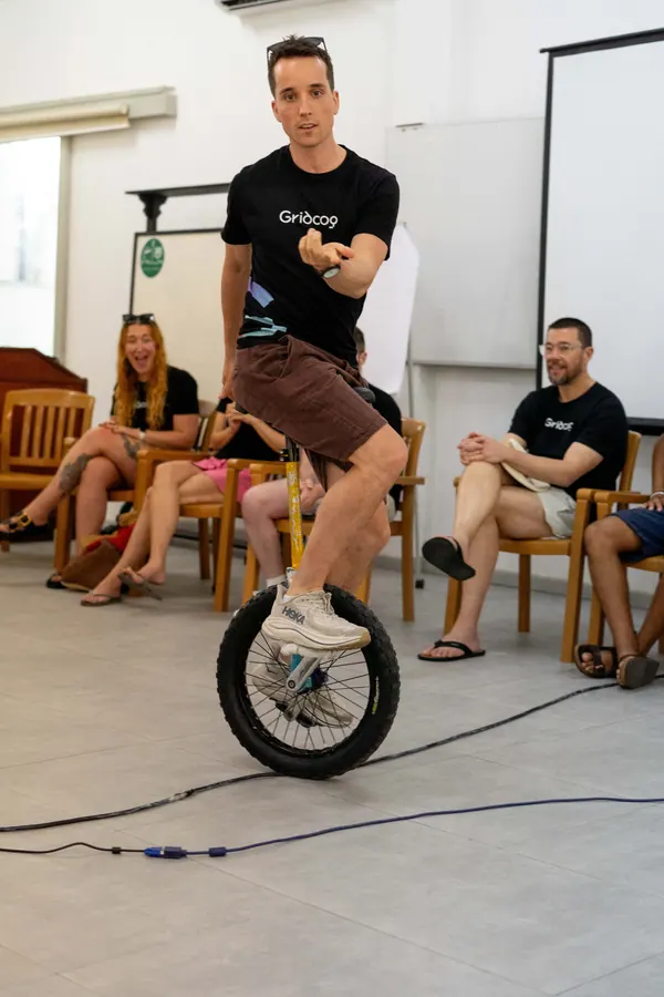 A Gridcogger riding a unicycle while teammates watch and laugh