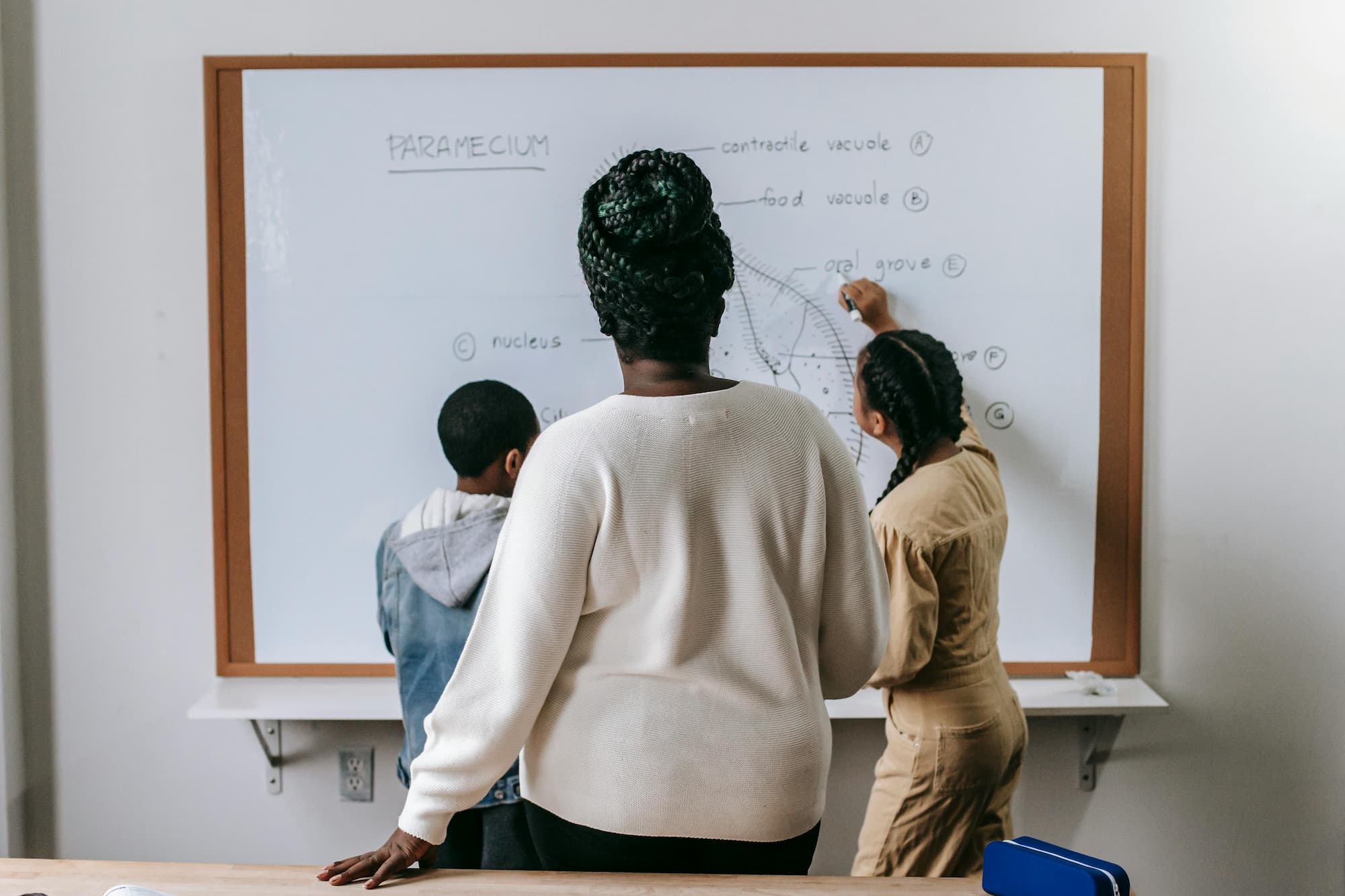 A teacher in a school classroom standing in front of a chalkboard filled with handwritten mathematics, arms crossed, looking at the camera.