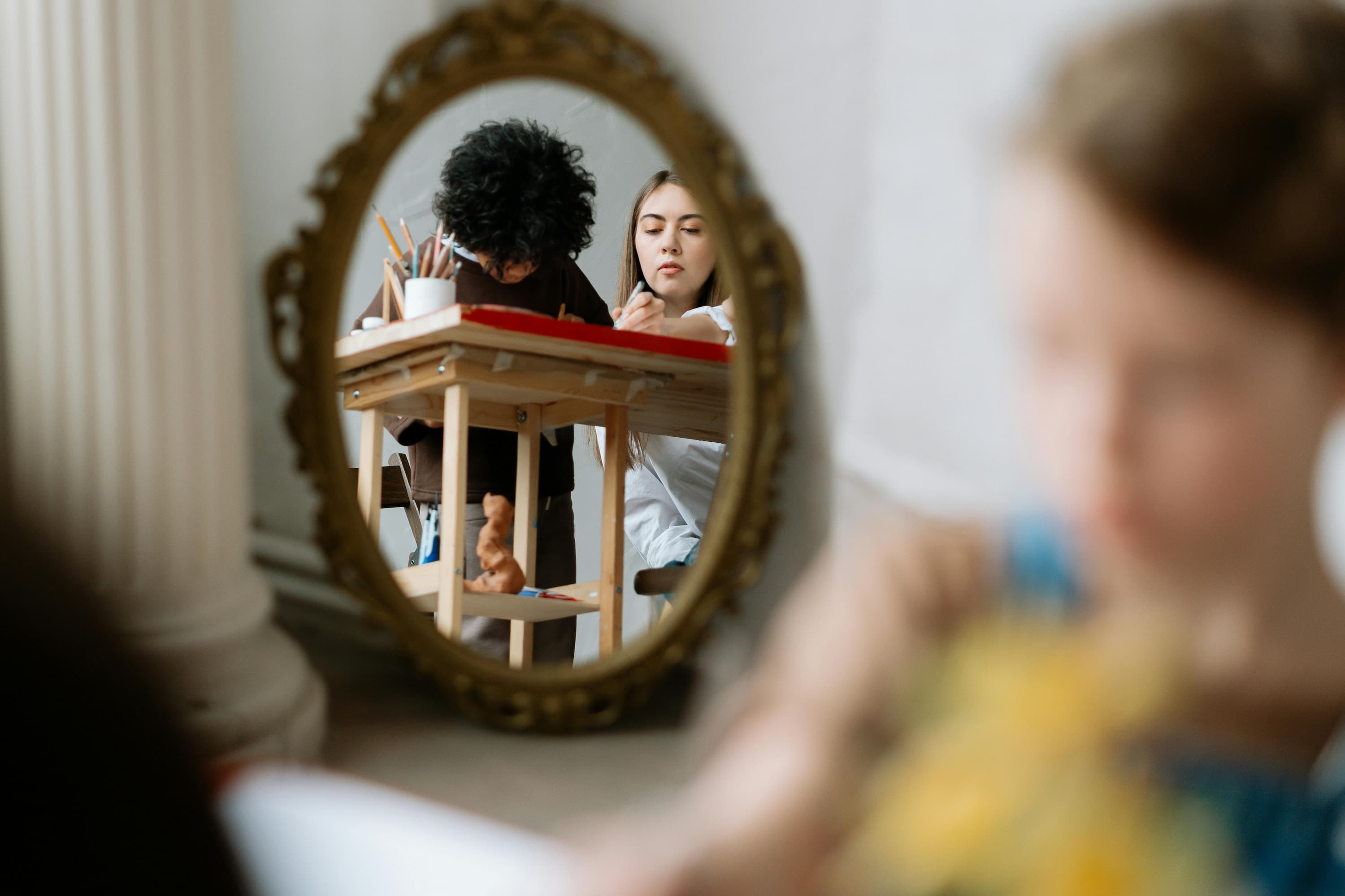 An ornate gold-framed oval mirror reflecting a young woman and a child working together at a wooden art table, with the child’s head out of focus in the foreground.