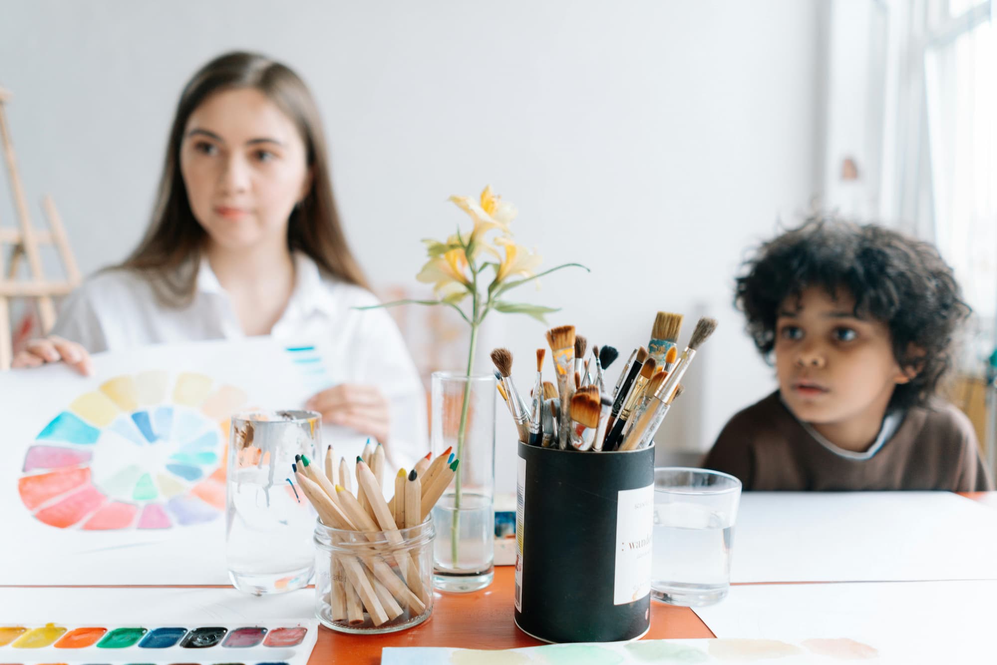 Paintbrushes, colored pencils, and a watercolor palette in the foreground of a bright art studio, with a teacher holding a color wheel and a child seated across the table, both out of focus in the background.