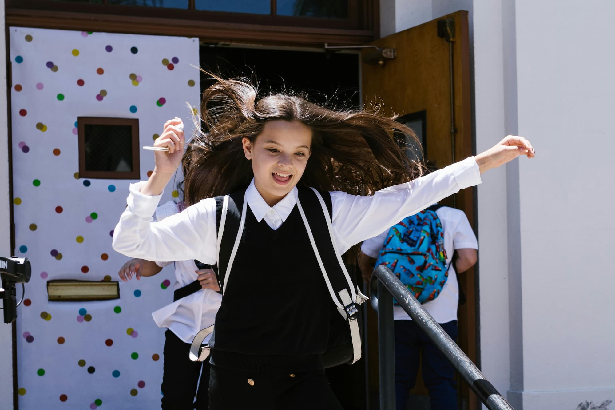 A middle-school student in uniform mid-stride, arms outstretched and hair flying, leaving the school’s front doors.