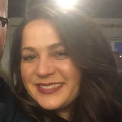 Smiling woman with long brown hair and dark lipstick in a dimly lit indoor setting.