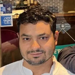 Close-up of a man with short curly hair and a trimmed beard, wearing a white shirt, sitting indoors.