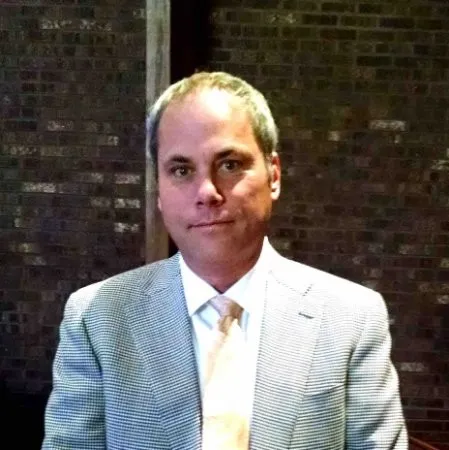 Man in a light checkered suit jacket and white shirt with a light-colored tie, sitting against a dark brick wall background.