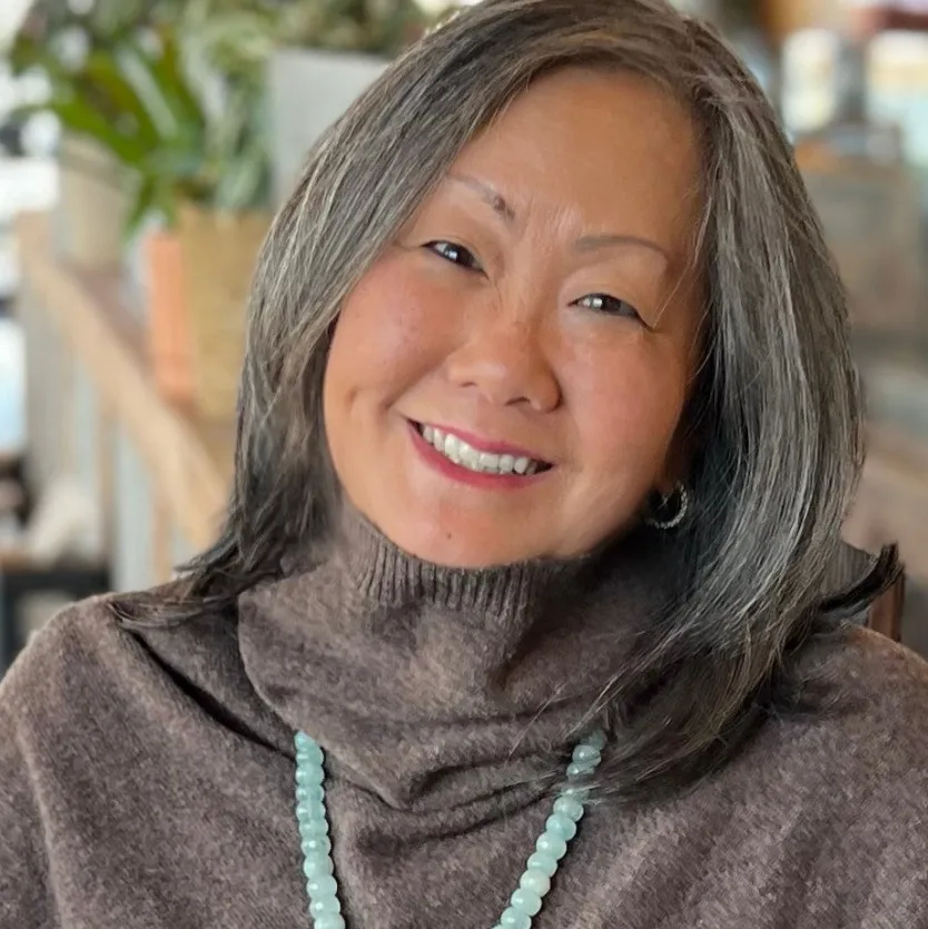 Smiling woman with gray hair wearing a brown turtleneck sweater and a light blue bead necklace.