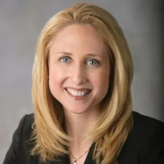 Smiling woman with shoulder-length blonde hair wearing a black jacket against a gray background.