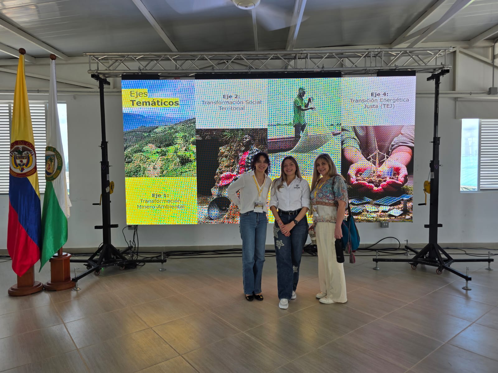 Tres mujeres de pie posando frente a una pantalla con la presentación de ejes temáticos; a un costado se observan banderas institucionales en el espacio del evento.