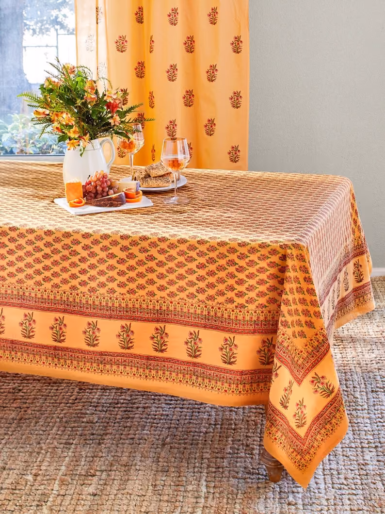 May include: A yellow and orange floral tablecloth with a red border. The tablecloth is laid on a woven rug and has a plate of fruit and two wine glasses on top.