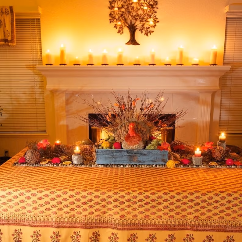 May include: A table with a yellow and orange patterned tablecloth. A blue wooden box with a fall floral arrangement is on the table. The arrangement includes a turkey figurine, fall leaves, and pine cones. There are candles on the table and a fireplace in the background.