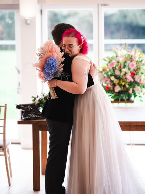 Bride with bright pink hair and glasses hugging a person in a black suit while holding a pink and blue floral bouquet.