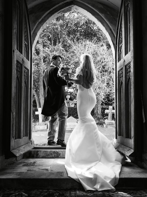 Black and white photo of a bride in a fitted gown holding a bouquet and stepping outside an arched doorway with a groom in a suit looking back and smiling.