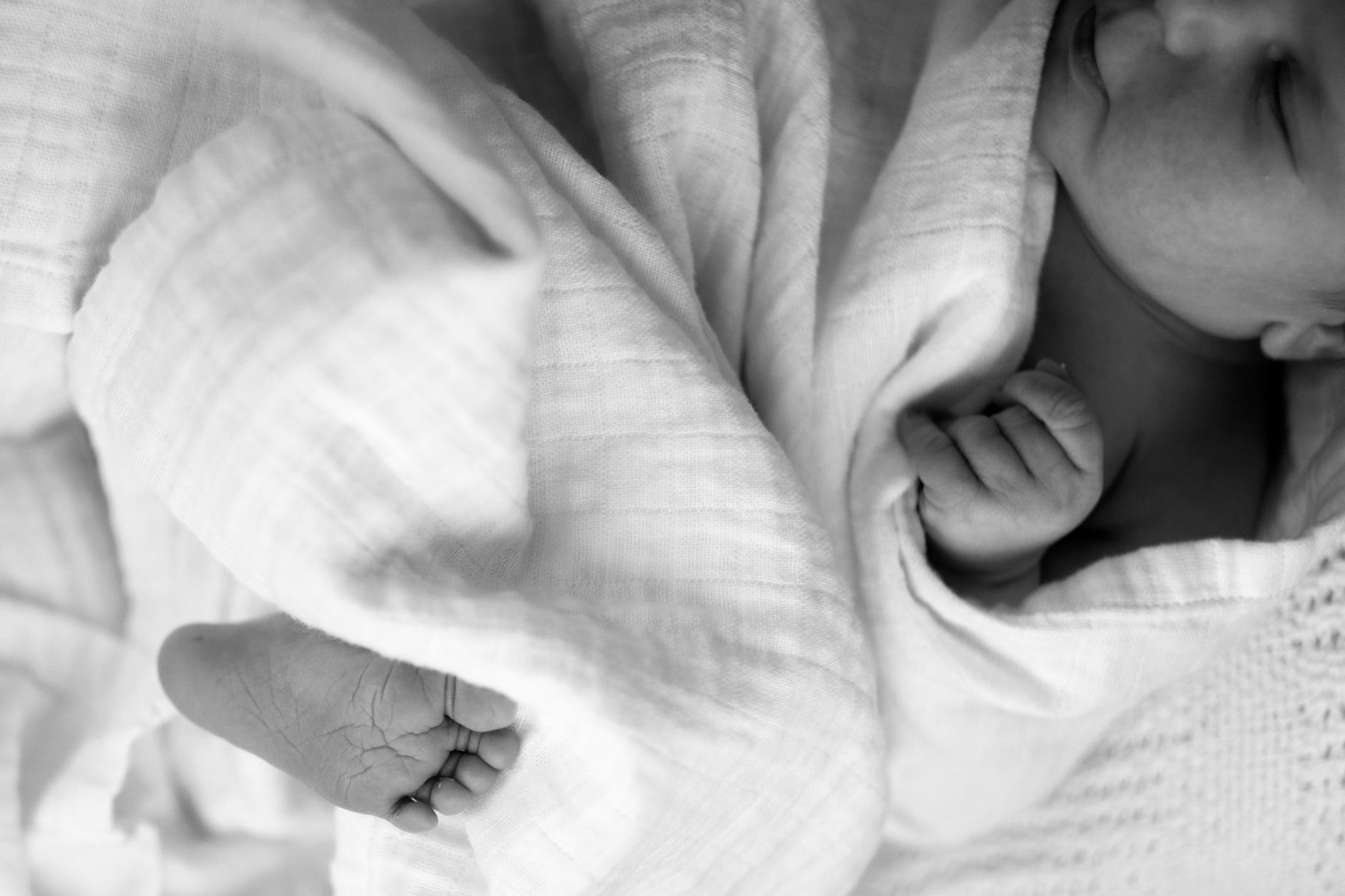 Black and white close-up of a newborn baby wrapped in a soft blanket showing its tiny hand and foot.
