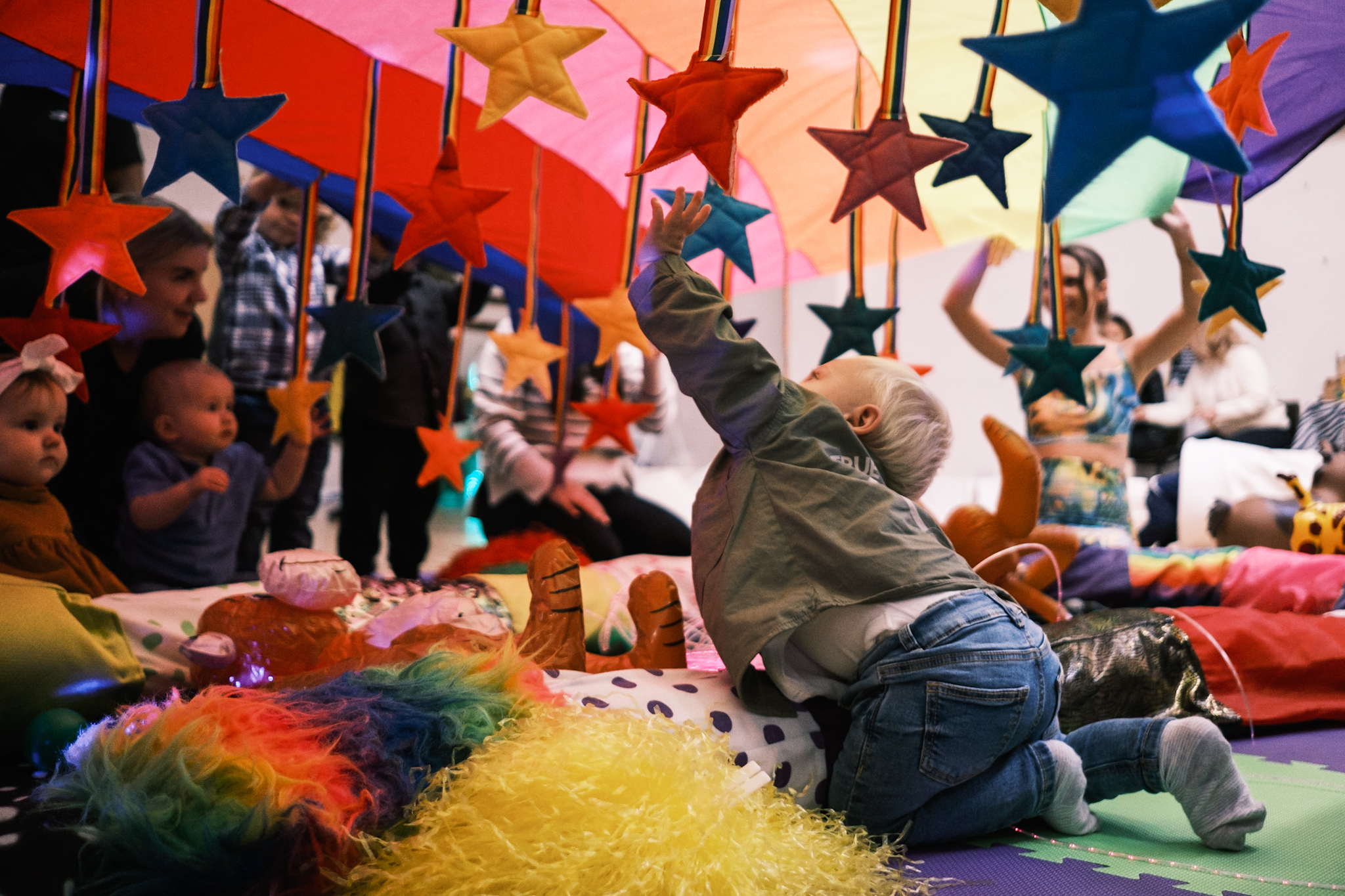 A young child reaches up towards hanging colorful star-shaped decorations under a rainbow-colored parachute in a playful group setting with other children and adults.