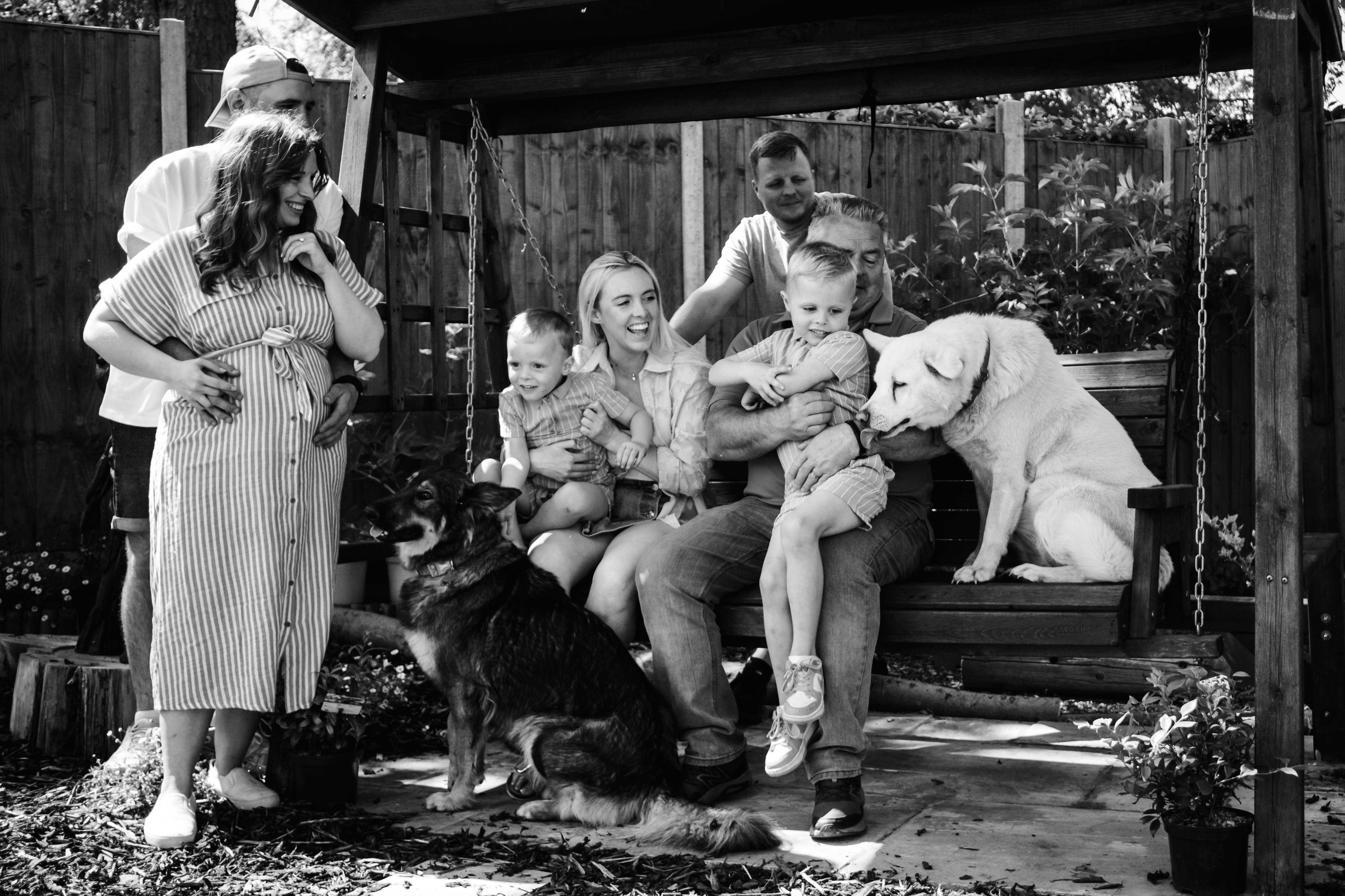 Black and white photo of a multi-generation family with two young boys and two dogs enjoying time together on a garden swing and standing nearby.