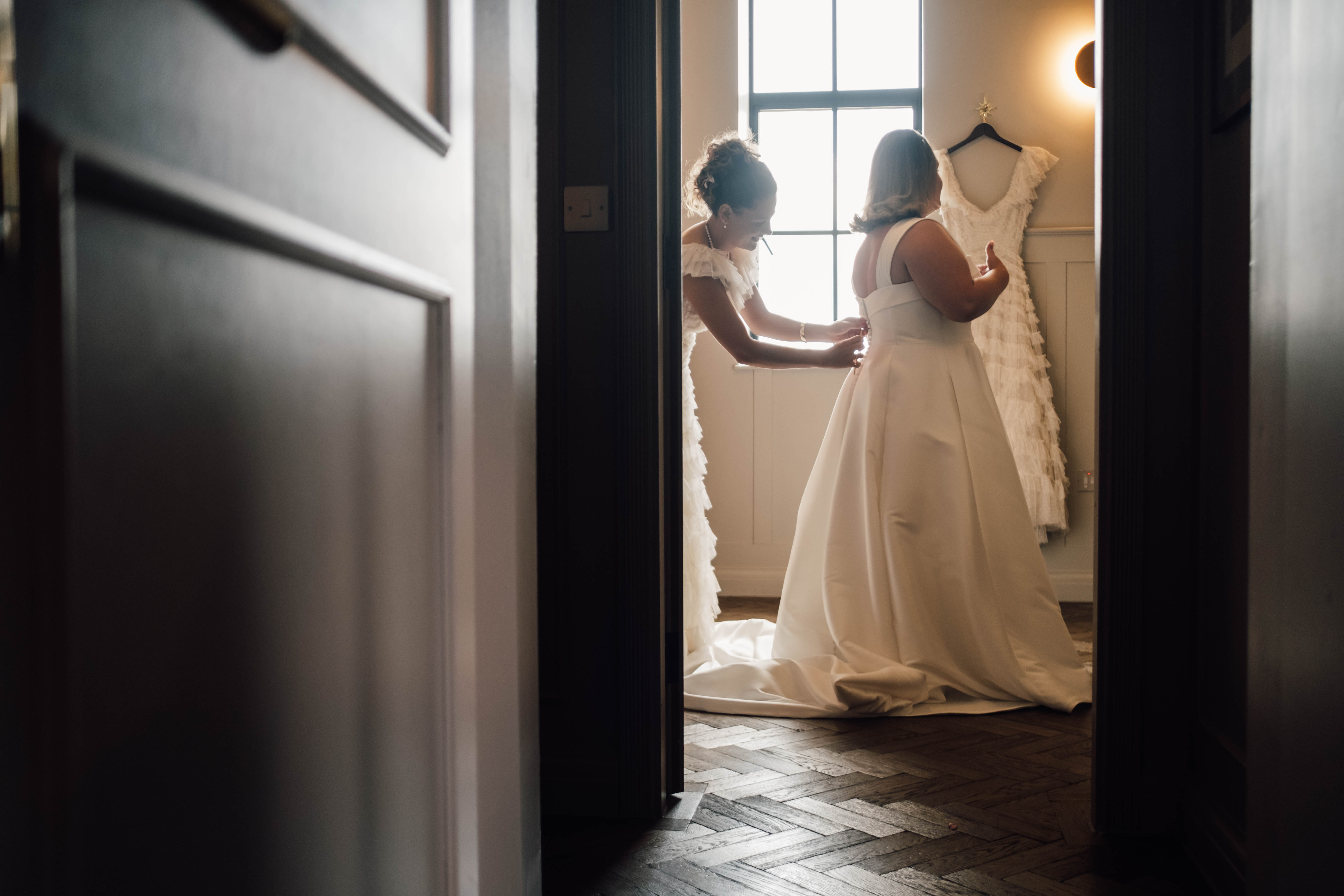 Woman in a bridal gown being helped with buttoning the back of her dress by another woman in a softly lit room.