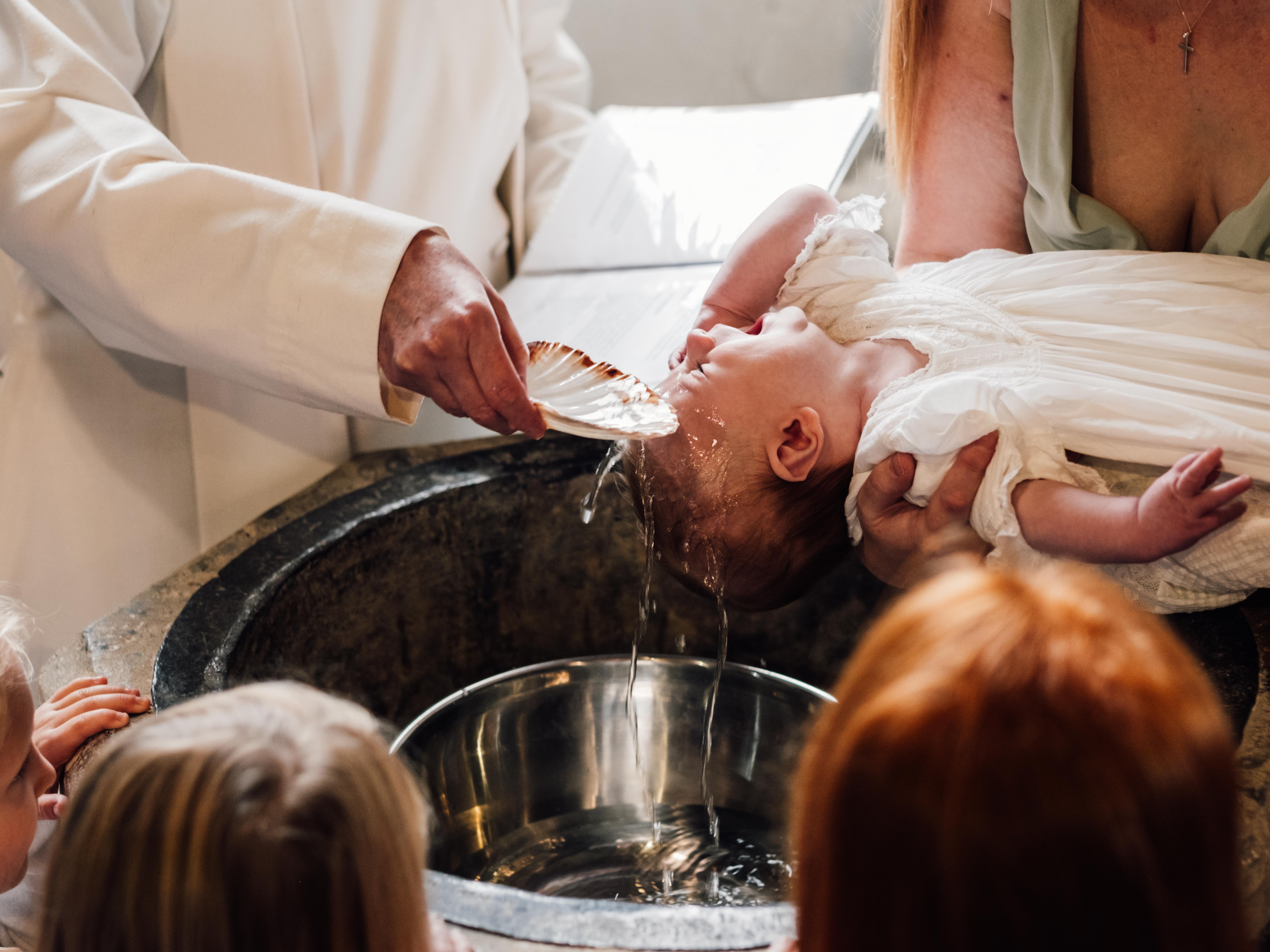 Groom in patterned vest lifting bride in white wedding dress under shaded structure with greenery and sunset in background.