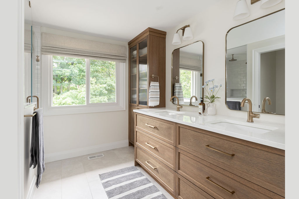 Renovated bathroom with warm oak double vanity, white quartz countertop, brushed brass fixtures, and a glass-front linen cabinet with natural light