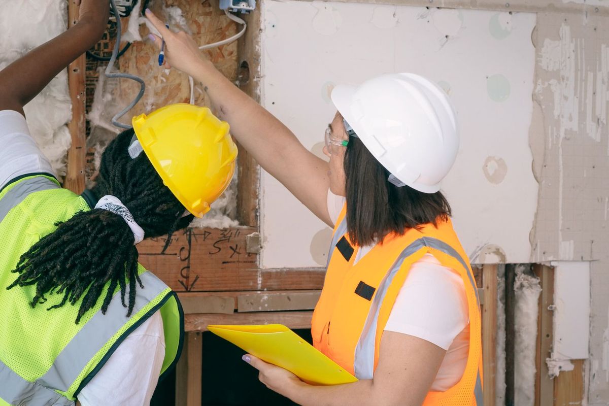 Two renovation professionals in hard hats and safety vests reviewing old insulation and wiring behind an opened wall, one holding a clipboard