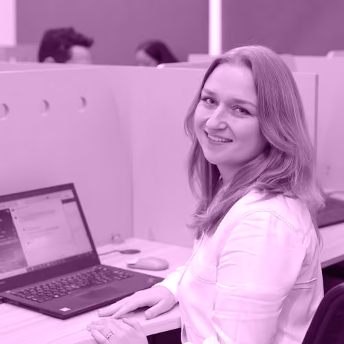 Smiling woman sitting at a desk with a laptop in an open office workspace.