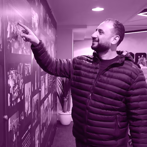 Man in a puffy jacket smiling and pointing at images on a wall display indoors.