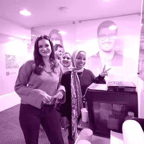 Four smiling young adults in casual clothes posing in a modern office space near a coffee machine.