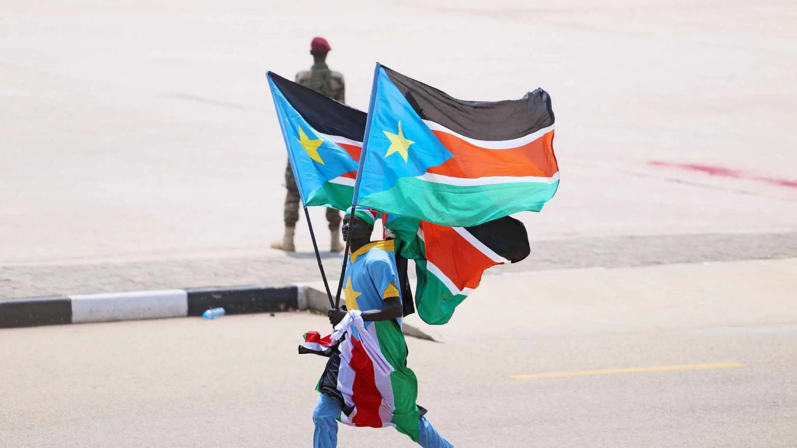 A man carrying two South Sudanese flags in Juba, with a soldier standing in the background