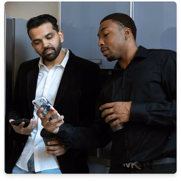 Two men in business casual attire standing together, one showing a phone screen to the other while holding coffee cups.