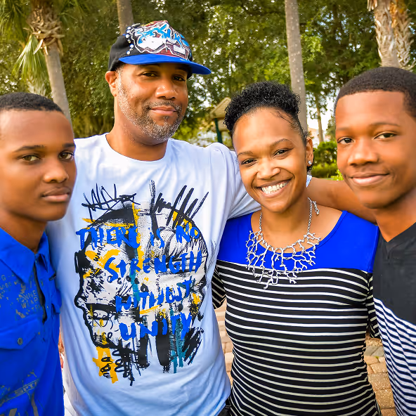 Smiling family of four standing closely outdoors with trees in the background.