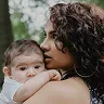 Curly-haired woman gently holding and kissing a baby resting on her shoulder outdoors.