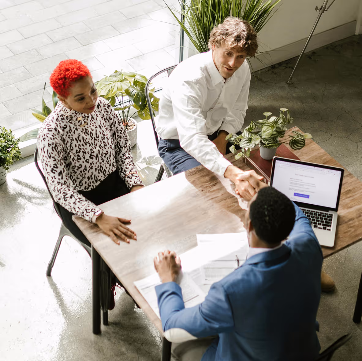 Two people sitting at a table shake hands with a man in a blue suit during a meeting in a bright office with indoor plants.
