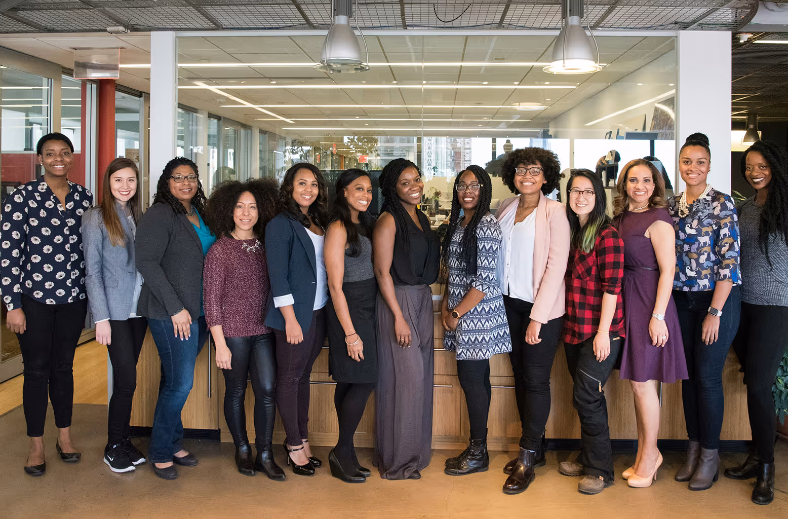 Group of thirteen diverse professional women standing indoors in an office setting, smiling at the camera.
