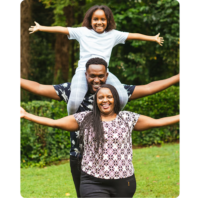 Smiling family of three with child sitting on father's shoulders, all with arms outstretched in a green outdoor setting.