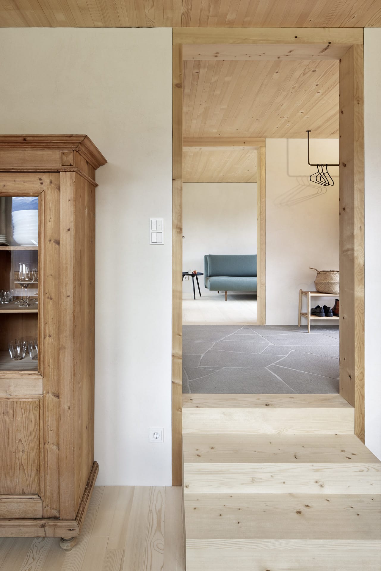 Küchenschank mit Weingläsern neben einer dreistufigen Treppe, welche in den Eingangsbereich des Hauses führt. Im Hintergrund ist ein Teil der Couch vom Wohnzimmer zu erkennen.