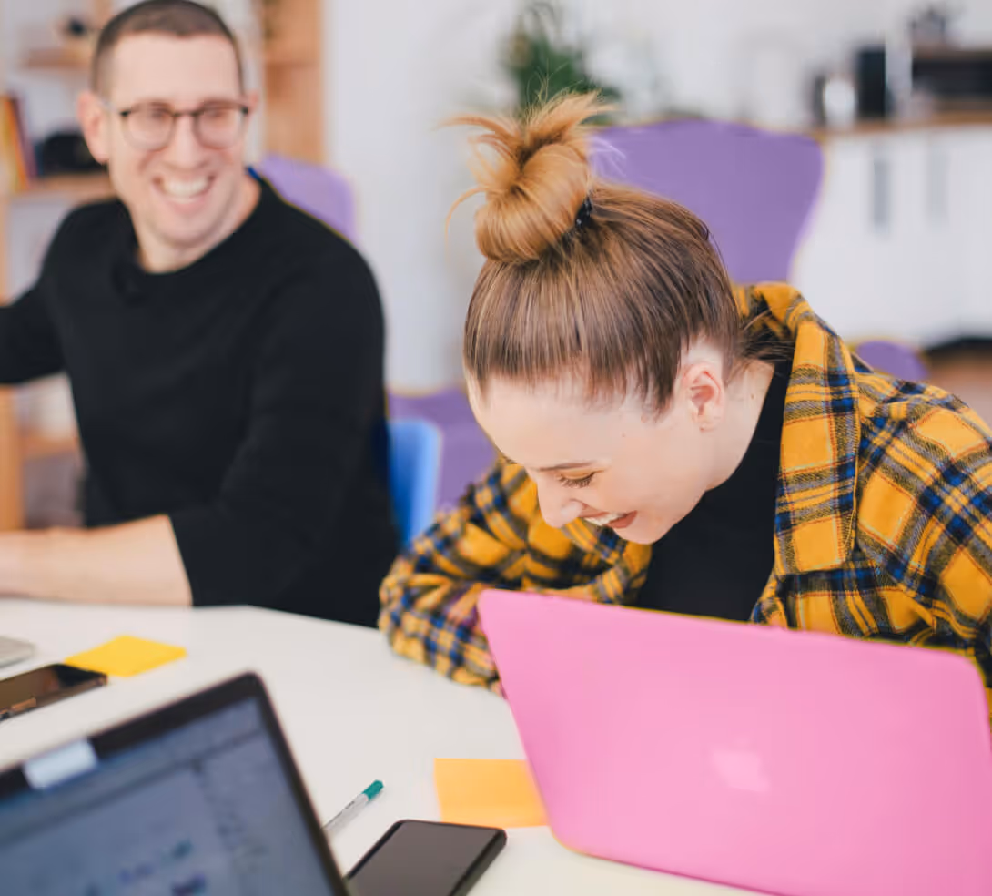 A man and woman sit at a table, each using a laptop, engaged in conversation or work.