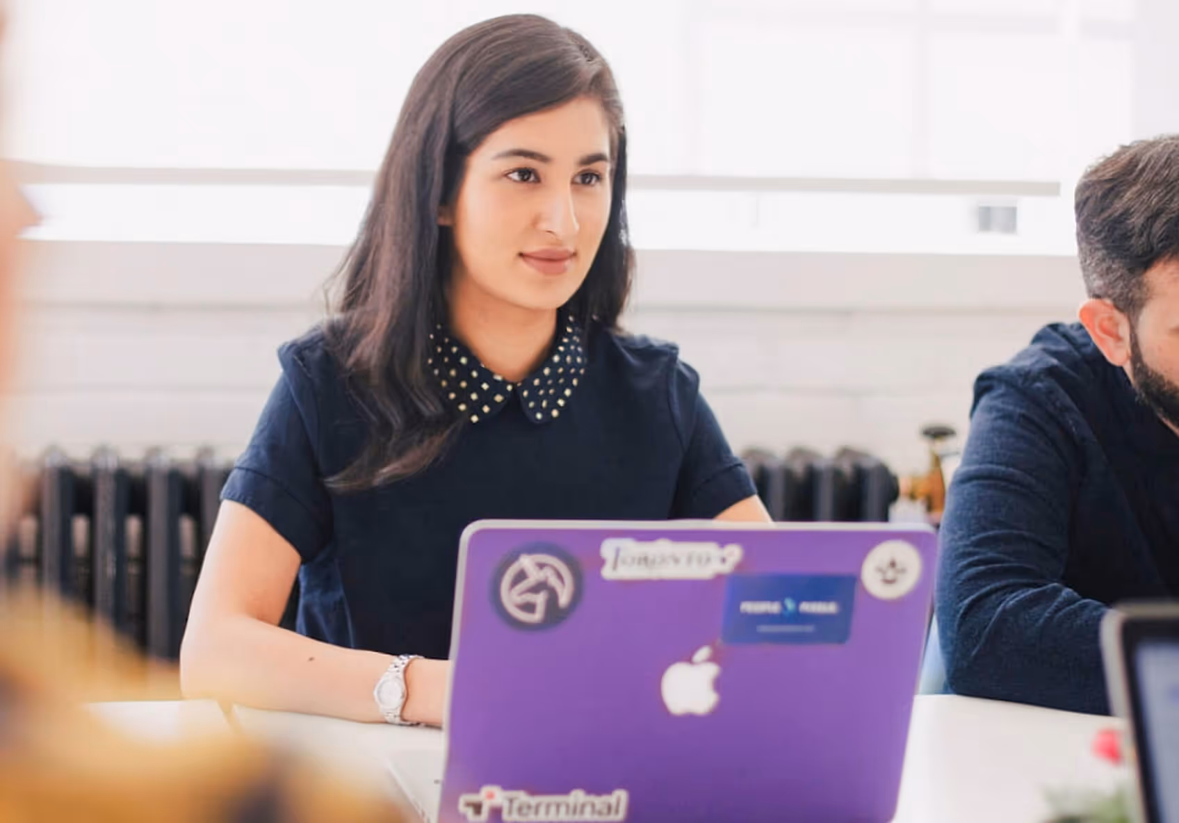 A woman and a man sit at a table with laptops, engaged in conversation in a professional office setting.