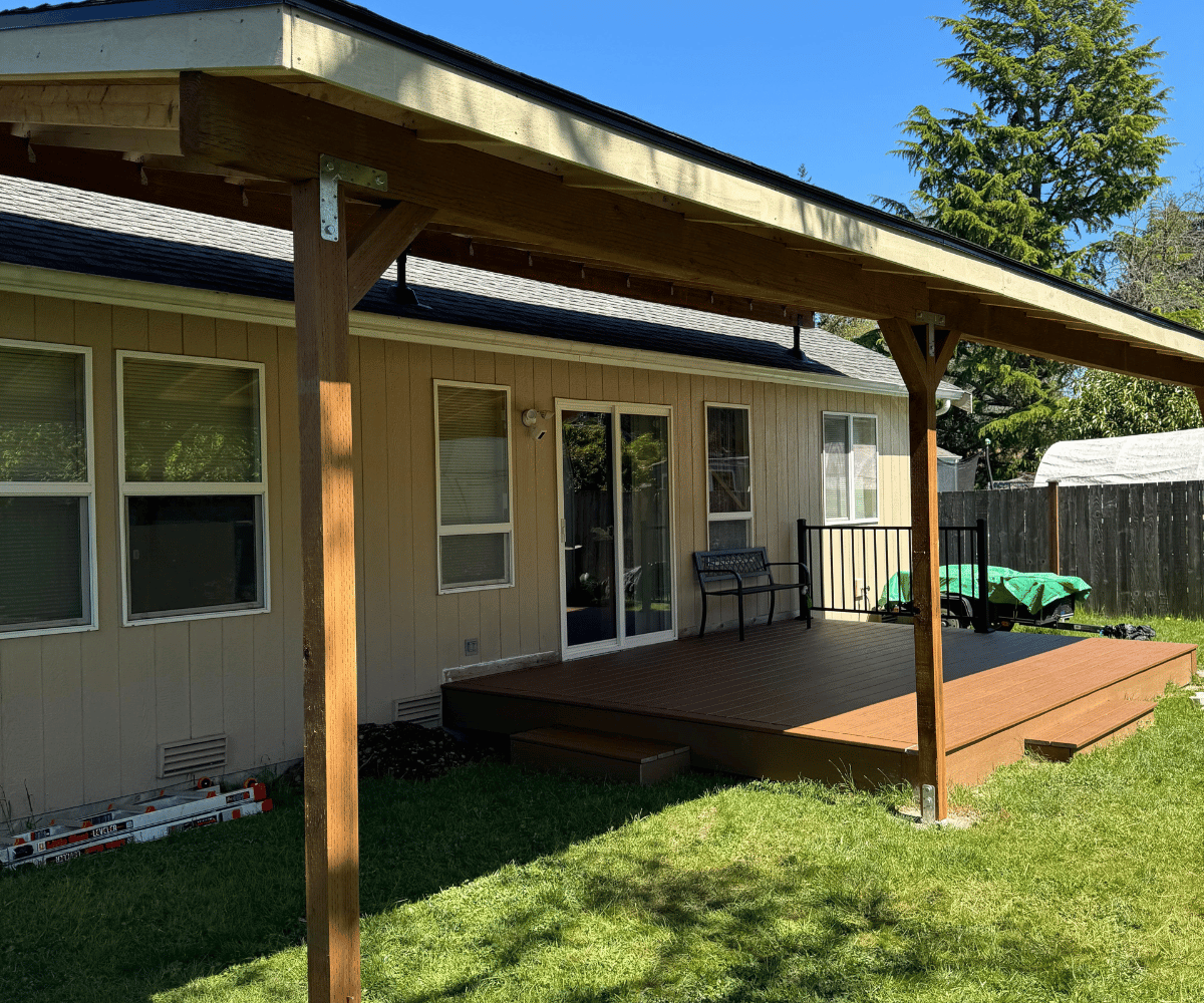 Back patio with wooden deck and pergola attached to beige house under clear blue sky.