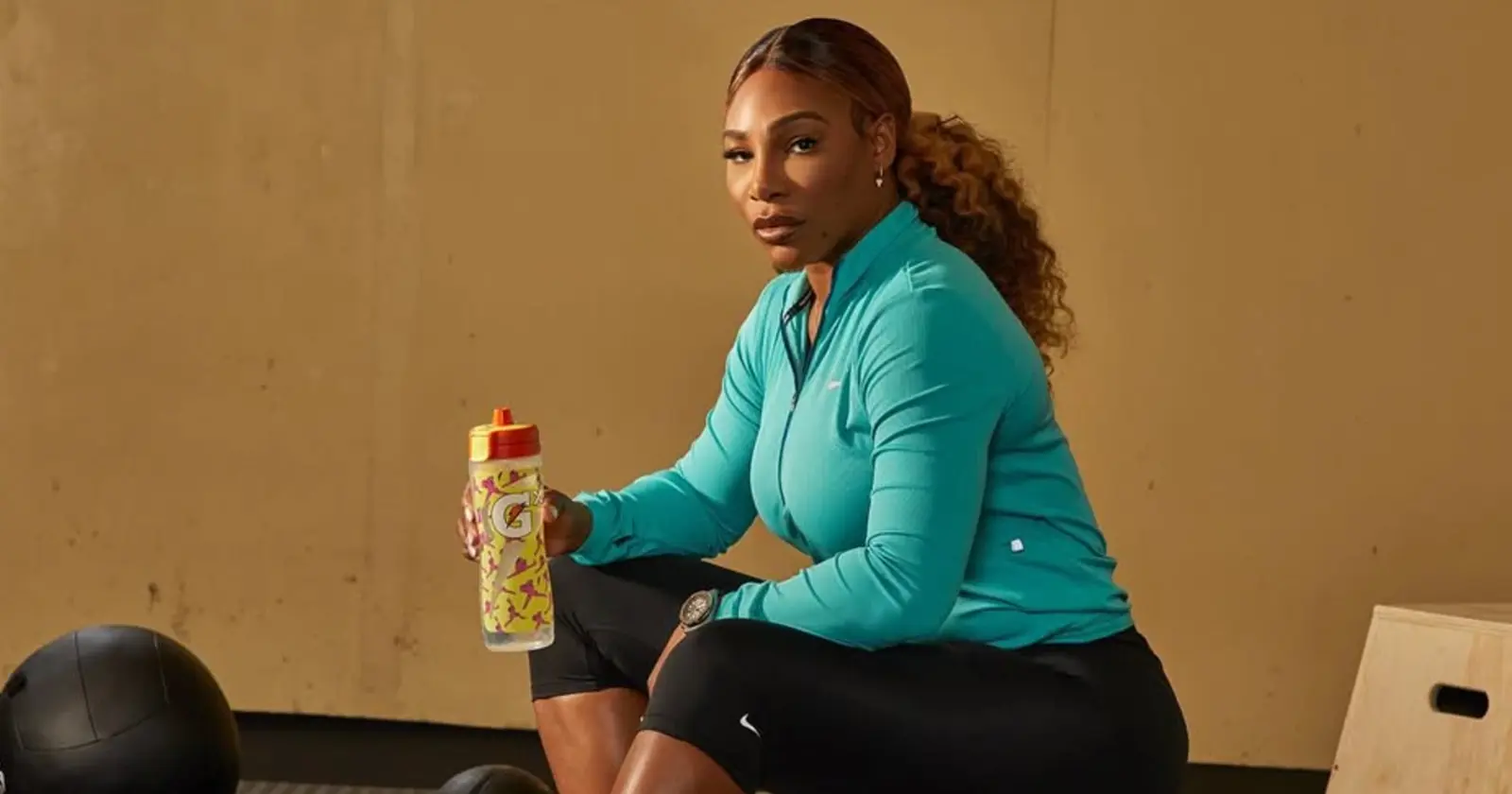 Serena Williams seated in a gym wearing a turquoise athletic jacket, holding a Gatorade bottle during training.