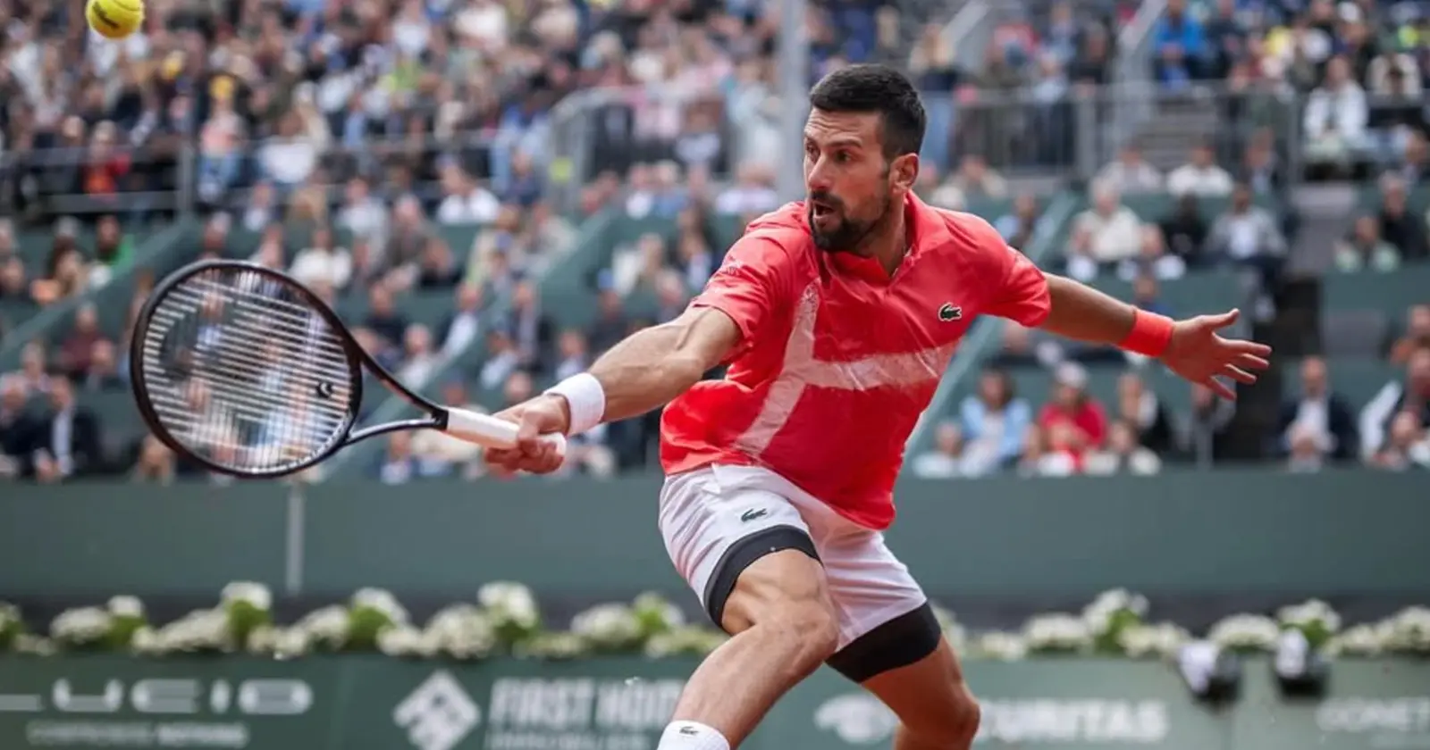 Novak Djokovic stretches wide for a forehand return during a Grand Slam tennis match, wearing a red Lacoste shirt on a packed court.