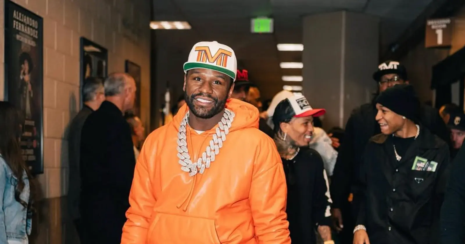 Floyd Mayweather wearing an orange hoodie and large diamond chain smiles while walking with his team.