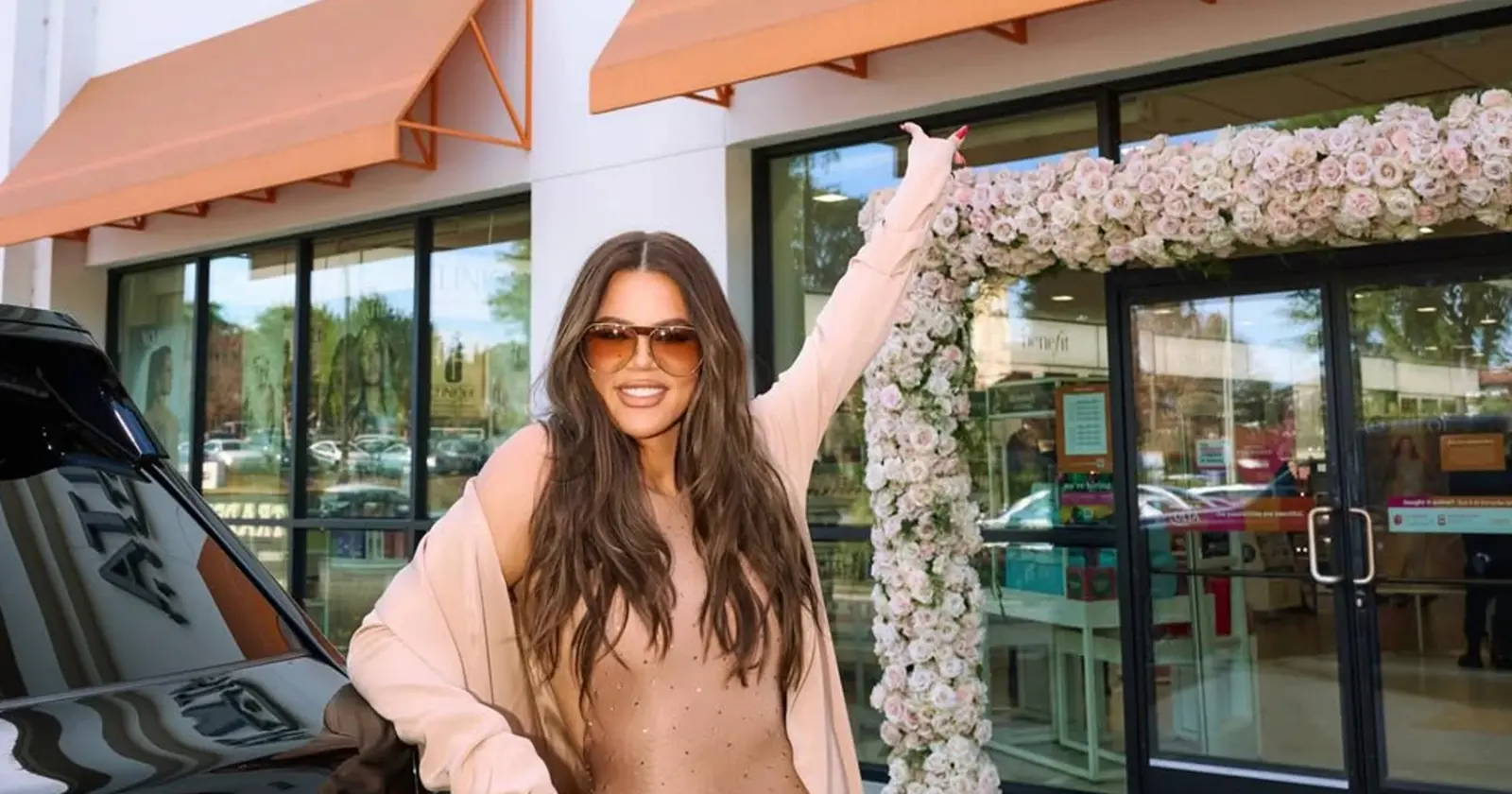 Khloé Kardashian raises her hand outside a floral-decorated storefront during a promotional event.