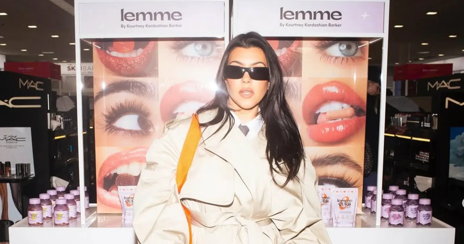 Kourtney Kardashian in sunglasses and a beige trench coat poses in front of a Lemme product display at a beauty retailer.