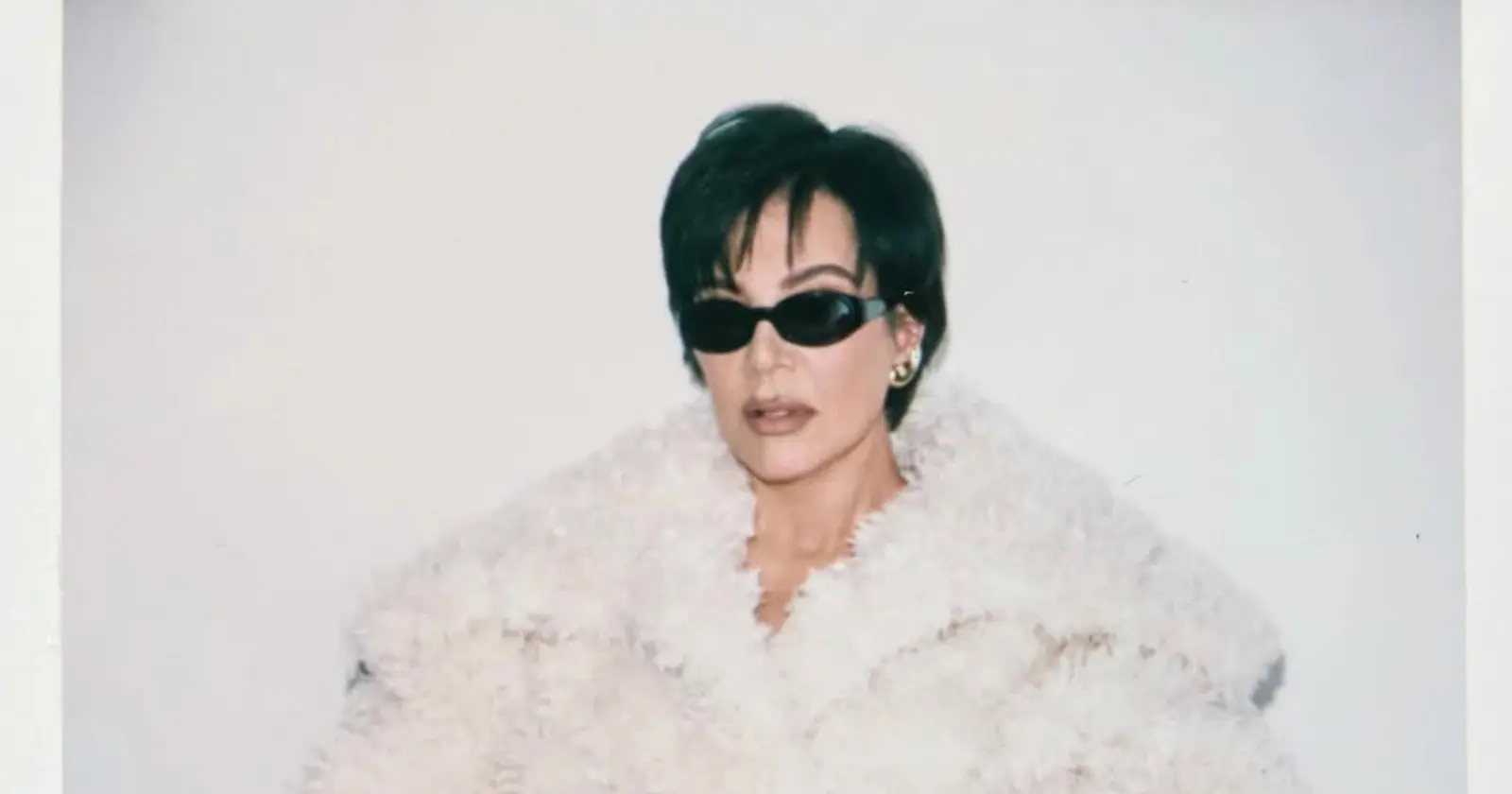 Kris Jenner wears a dramatic white fur coat with dark sunglasses in a striking fashion portrait.