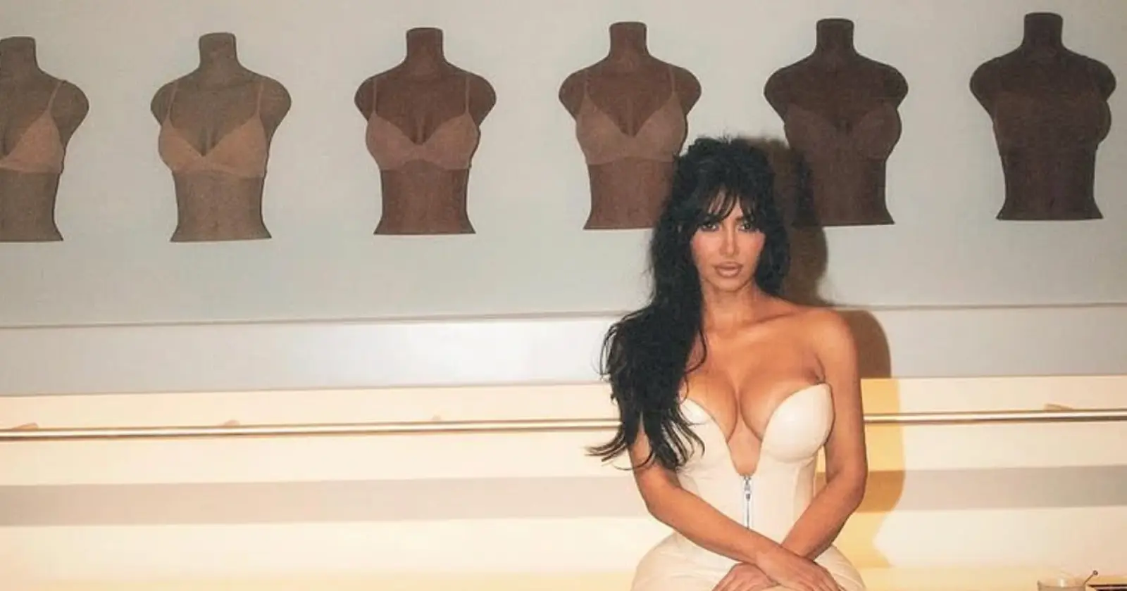 Kim Kardashian poses in a cream corset dress in front of Skims mannequin displays highlighting her shapewear brand.