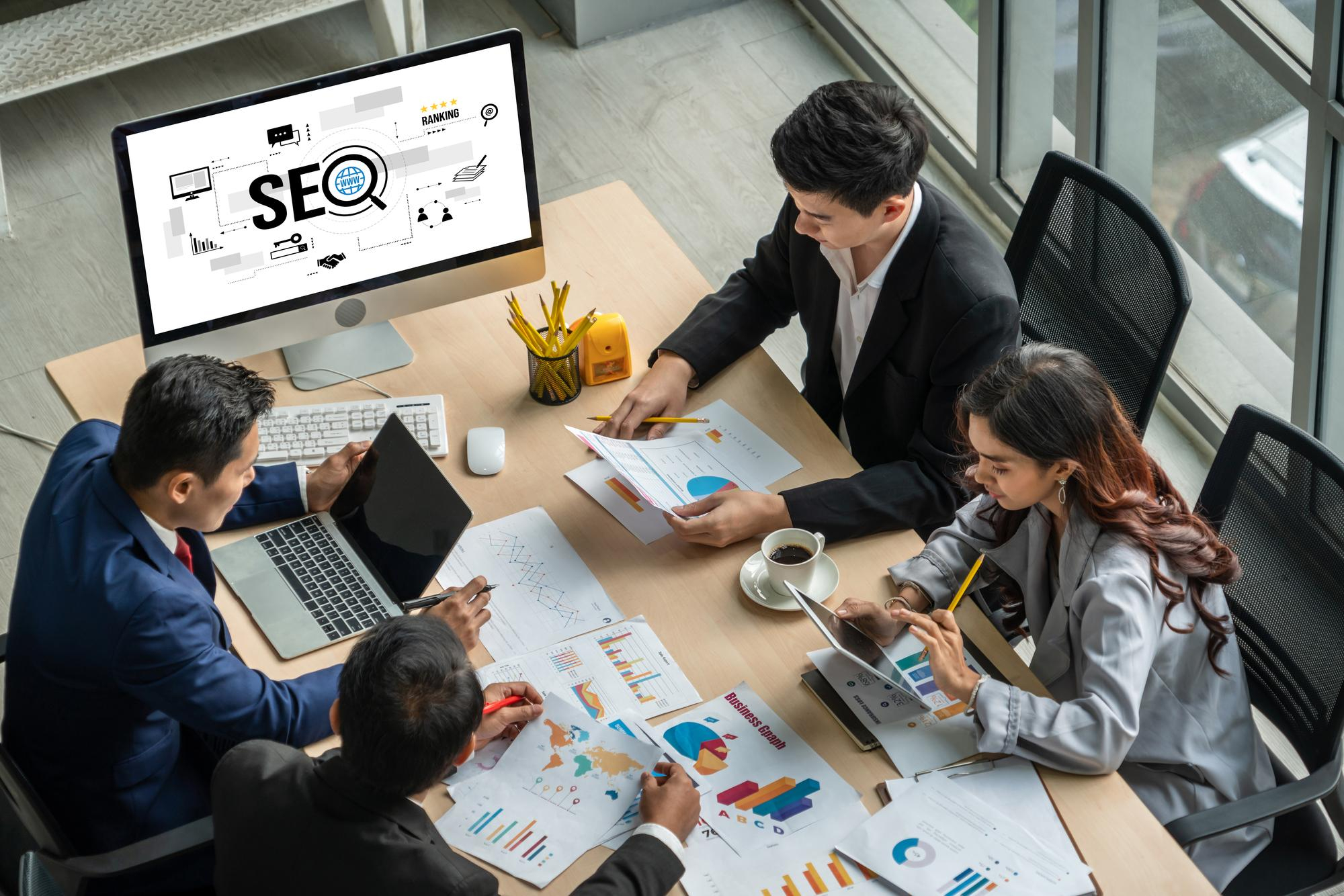 The marketing team is reviewing SEO strategy charts and reports around a boardroom table with an SEO monitor display in the background.