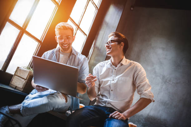 Two smiling male colleagues collaborating on laptop in modern office workspace with large windows.